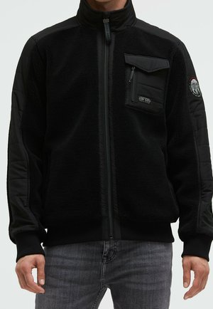 Mann trägt eine schwarze Fleecejacke mit Frontreißverschluss, Reißverschlusstasche auf der Brust und Aufnäher am oberen linken Ärmel, kombiniert mit grauer Jeans.