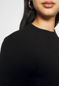 Svart stickad tröja med rund halsringning och ribbad struktur, visad från axel till haka. Stora hoops örhängen synliga.