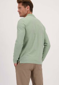 Lichtgroene polo met lange mouwen en een gestructureerd patroon, zijventen en een contrasterende kraag, gecombineerd met beige broeken.