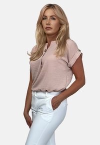 Jasnoróżowa bluzka z krótkimi, podwiniętymi rękawami i subtelnie wyciętym dekoltem w kształcie V, w połączeniu z białymi dopasowanymi spodniami. Gładka faktura, detal w postaci złotego guzika.