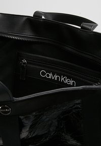 Czarna torba na ramię z połyskującą, teksturowaną powierzchnią. Wnętrze zawiera kieszeń na zamek błyskawiczny z białą metką wyświetlającą "Calvin Klein."