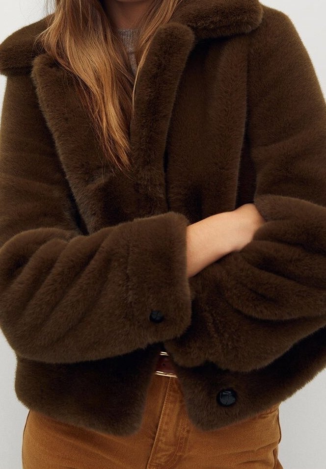 Femme portant une veste en fausse fourrure pelucheuse marron foncé avec de grands revers et des boutons noirs, sur un pantalon beige, bras croisés à la taille.