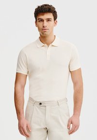 Beżowa koszulka polo z kołnierzykiem i trzema guzikami, wykonana z mi�ękkiego materiału. Noszona z jasnobeżowymi spodniami, z widocznym guzikiem i fałdami.