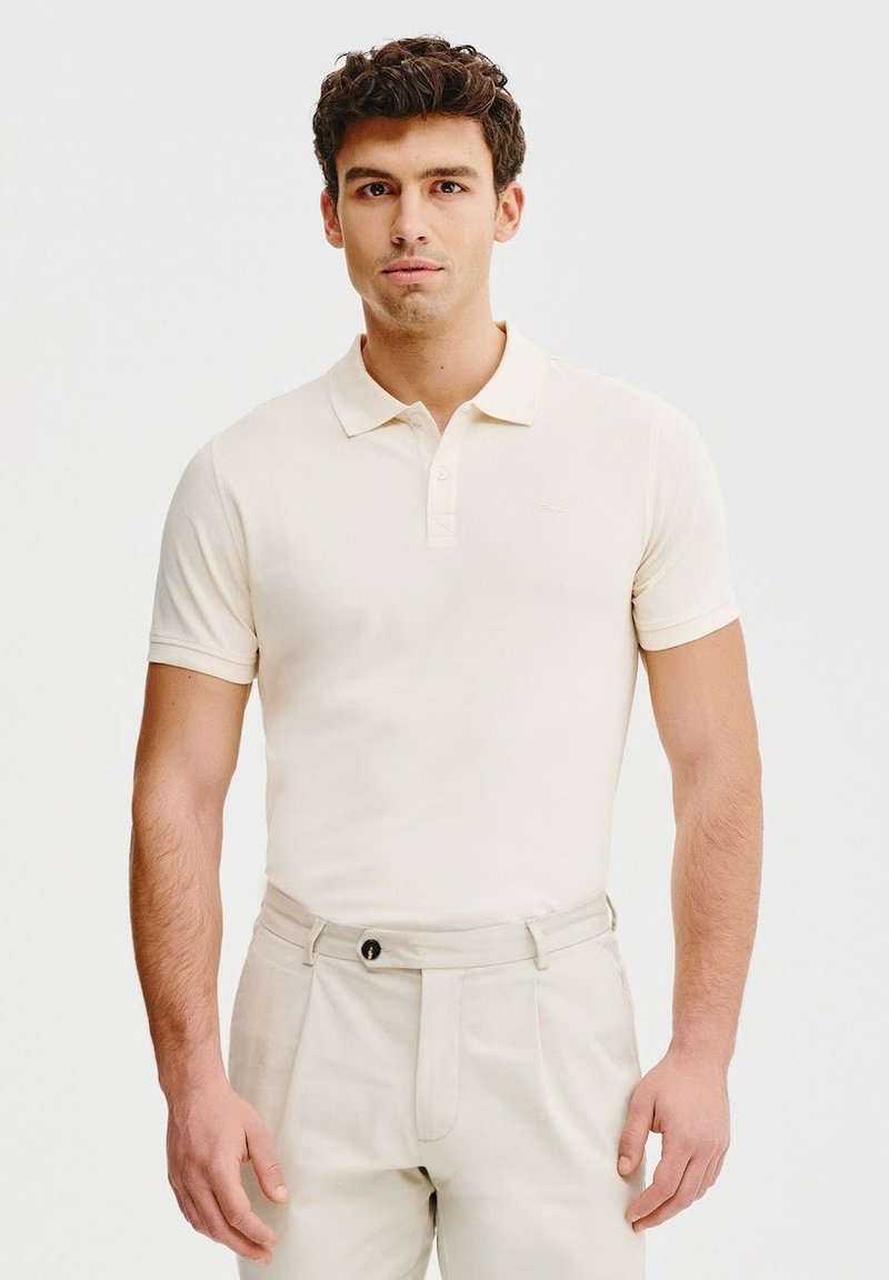 Beżowa koszulka polo z kołnierzykiem i trzema guzikami, wykonana z miękkiego materiału. Noszona z jasnobeżowymi spodniami, z widocznym guzikiem i fałdami.