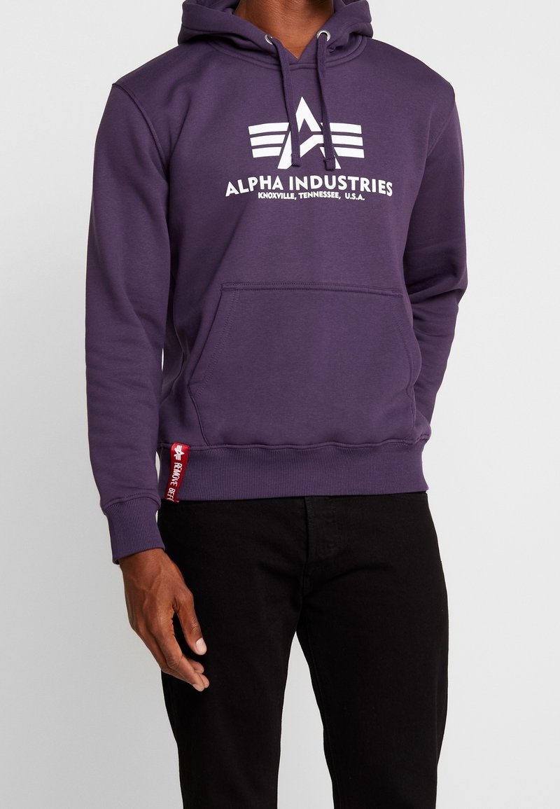 Homme portant un sweat à capuche violet Alpha Industries avec poche avant et pantalon noir, debout devant un fond uni.