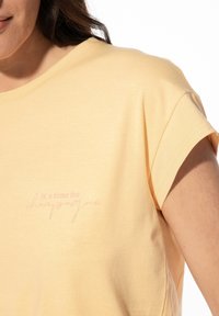 Lichtgele t-shirt gemaakt van zachte stof, met opgerolde mouwen en een roze bedrukte slogan: "het is tijd voor champagne" op de voorkant.