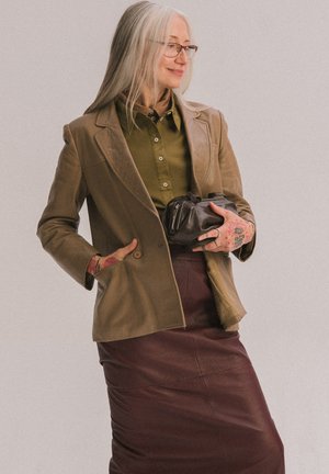 Femme âgée aux longs cheveux gris, portant des lunettes et des tatouages, vêtue d'une veste en cuir marron et d'une jupe, tenant une pochette noire, souriant de côté.