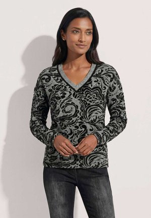 Femme portant un pull à manches longues noir et gris à motifs, col en V, et un jean foncé, debout, mains jointes de manière détendue.