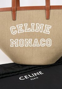 Sac cabas en toile beige avec des poignées en cuir marron et un texte blanc "CELINE MONACO", posé sur un tissu noir avec un "CELINE PARIS" blanc.