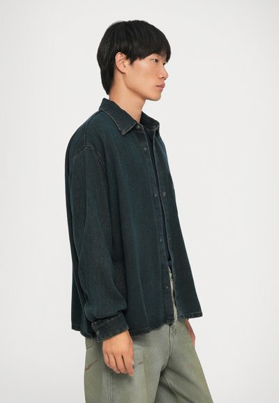Overdimensjonert mørkegrønn denimskjorte med knapper foran, krage og lange ermer. Brukt over en svart topp, sammen med lysegrønne jeans.