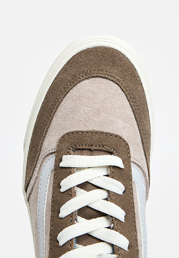 Baskets en daim de couleur marron et grise, avec une semelle en caoutchouc blanc et des lacets beige. Des coutures horizontales et un motif texturé subtil sont visibles.