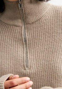 Beige vetoketjupaita, jossa on ribbikuviointi ja korkea kaulus. Varustettu hopeisella vetoketjulla ja pidennetyillä hihoilla, jotka korostavat lämmintä kangasta.