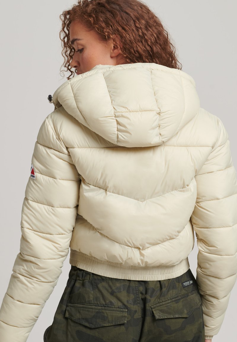 Giacca a vento beige con cappuccio, design trapuntato e orlo elasticizzato. Abbinata a pantaloni camouflage scuri. Presenta un logo sulla manica.