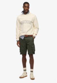 Sweat-shirt crème avec logo "SUPERDRY", shorts cargo olive, baskets beige et chaussettes montantes à rayures. Style streetwear décontracté.