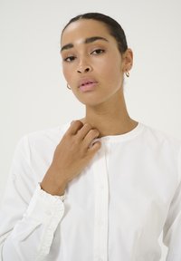 Chemise blanche à boutons avec un col arrondi et des poignets volants. Tissu en coton lisse ; modèle ajustant le col sur un fond neutre.