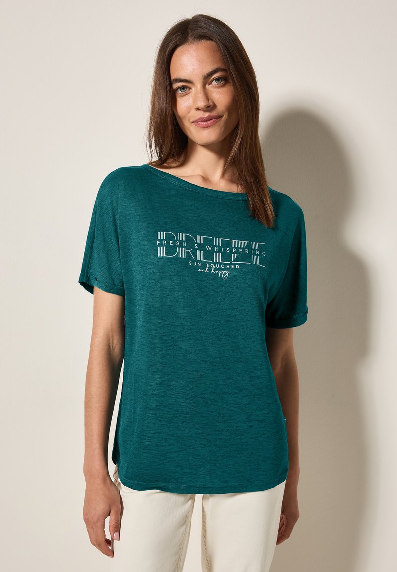Haut court teal à manches courtes avec une coupe décontractée, présentant un design de texte graphique blanc. Texture douce avec un col rond et des fentes latérales.