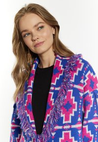 Cardigan tricotat cu un model geometric vibrant în nuanțe de albastru și roz, având un detaliu cu ciucuri pe guler. Confecționat dintr-un material moale, texturat.