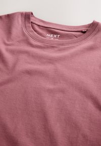 Camiseta de cuello redondo de color malva con etiqueta visible que muestra la marca "NEXT" y la talla mediana sobre un fondo claro.