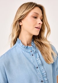 Light blue denim shirt with ruffled collar, front button closure, and short sleeves. Soft texture, casual design, and smooth finish.
