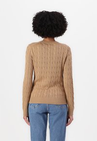 Un pull en maille torsadée couleur beige avec de longues manches, doté d'un col et d'un ourlet côtelés. Associé à un jean délavé, vu de dos.