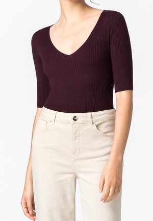Femme portant un haut côtelé violet foncé à col en V avec manches aux coudes et un pantalon taille haute blanc cassé avec poches avant.