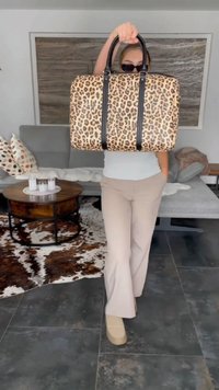 Pessoa a usar óculos de sol segura uma bolsa de padrão de leopardo à frente, de pé numa sala de estar moderna com um sofá e um tapete de pele de vaca.
