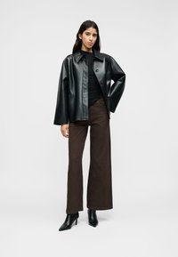 Schwarze Jacke aus Kunstleder mit Knopfleiste, getragen über einem schwarzen Oberteil, kombiniert mit braunen, weit geschnittenen Hosen und schwarzen Stiefeletten.