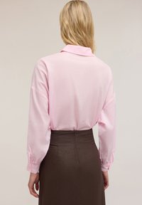 Blusa rosa chiaro con colletto, maniche lunghe e cadenti, e polsini con bottoni abbinata a una gonna marrone aderente con chiusura a zip.