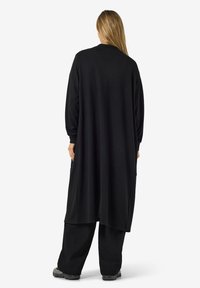 Lang svart cardigan med løs passform, rund hals, lange ermer og sidelommer. Myk tekstur, minimalistisk design.