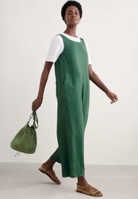 Žalias lino šokinėjimas su laisvu siluetu, turintis petnešas, šonines kišenes ir derinamas su paprasta balta marškinėliais bei rudi sandalai.