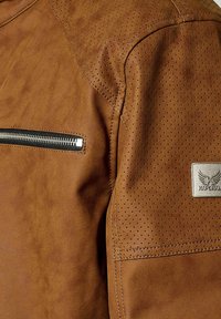 Veste en simili cuir marron avec des accents perforés aux épaules, poche zippée argentée et patch logo métallique sur la manche. Fini texturé.