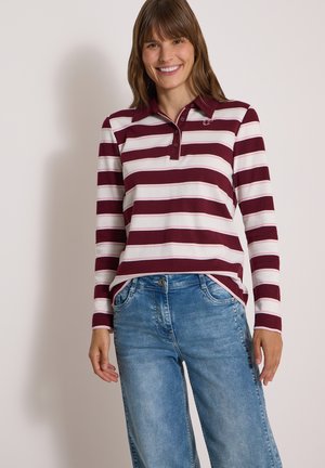 Frau lächelt, steht mit den Händen an den Seiten, trägt ein bordeauxrotes und weiß gestreiftes Langarm-Polohemd und blaue Jeans vor neutralem Hintergrund.