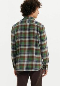 Camisa de franela a cuadros en verde, marrón y azul, con mangas largas y botones, que presenta un dobladillo curvado y detalle en el yugo trasero.