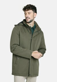 Hombre ajustando la manga de una chaqueta de invierno con capucha verde oliva, vistiendo pantalones beige y una camiseta verde oscura, contra un fondo blanco.