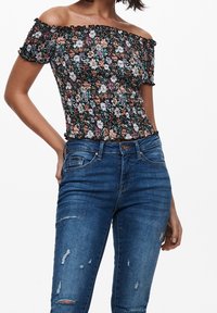 Olkalta paljastava kukkakuosiset paita, jossa on musta smokkikangas, jossa on valkoisia ja oransseja kukkia, yhdistettynä kulutettuihin sinisiin farkkuihin.