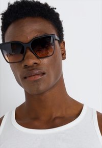 Black oversized sunglasses with gradient brown lenses, featuring a bold geometric shape and thick frames. Worn by a person in a white tank top.