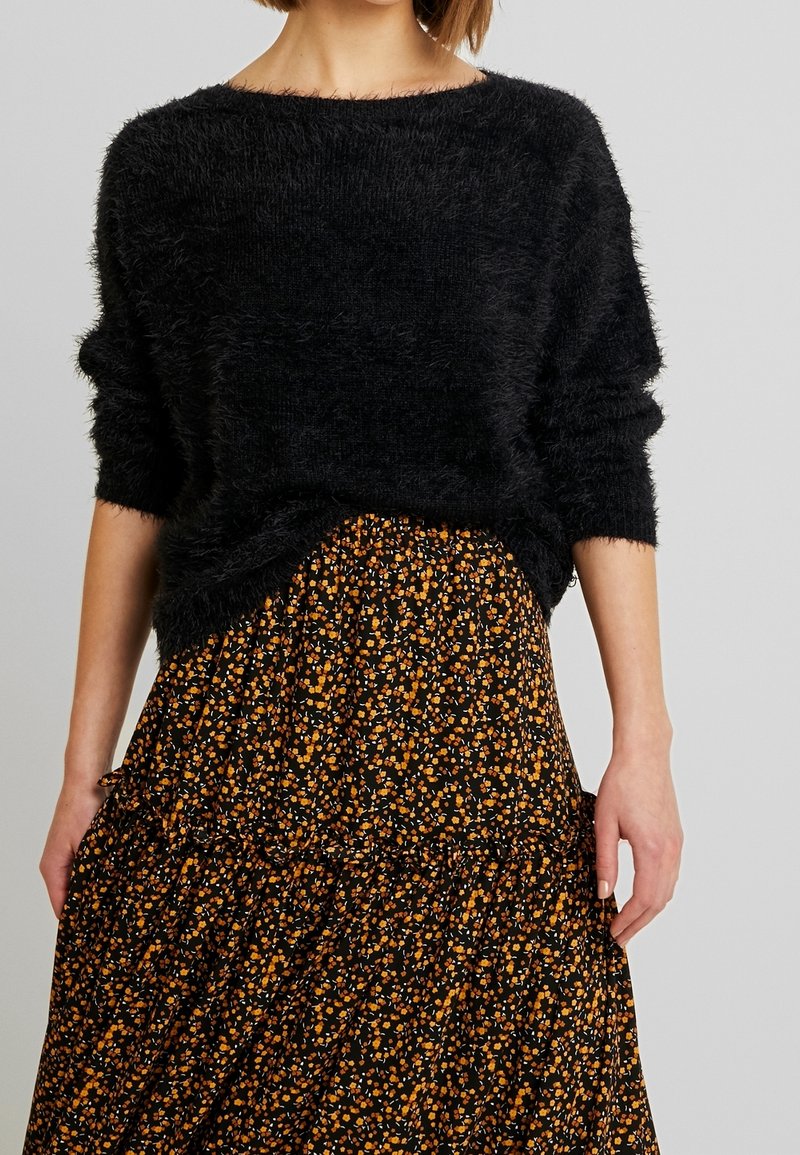 Schwarzer, flauschiger Strickpullover mit weitem Ausschnitt, kombiniert mit einem schwarzen Blumensrock, der kleine orange und weiße Blumen sowie gestufte Rüsche hat.