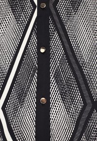 Tessuto con motivo geometrico in bianco e nero con forme a diamante, accentuato da una fila verticale di tre bottoni rotondi argentati riflettenti.