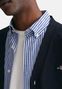 Marineblauer Cardigan über einem blau-weiß gestreiften Hemd mit Knöpfen, darunter ein weißes T-Shirt. Sichtbare Knopfdetails und Kragen.