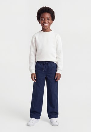 Enfant souriant debout, portant un t-shirt blanc à manches longues, un pantalon bleu marine avec cordon de serrage, et des baskets blanches sur un fond uni.
