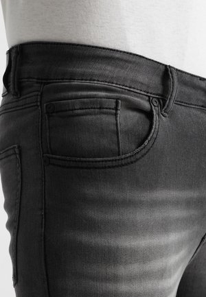 Close-up van een broekzak van zwarte spijkerbroek, met details van de stiksel en de textuur van de stof, gedragen door een persoon met een wit shirt.