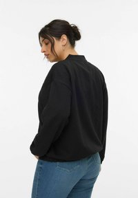 Giacca nera trapuntata con maniche lunghe e colletto alto, caratterizzata da una vestibilità comoda e vita elasticizzata, abbinata a jeans blu.