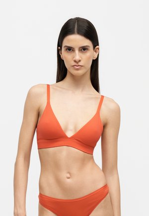 Mujer con cabello oscuro y liso que lleva un bikini de dos piezas naranja sobre un fondo claro y liso.