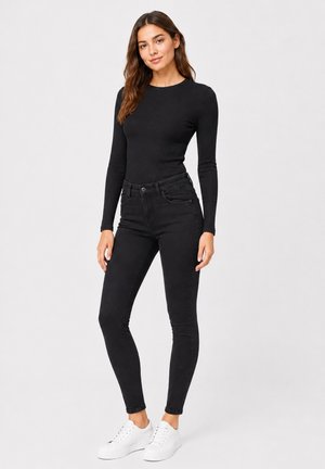 Kvinde stående iført en sort langærmet top, sorte skinny jeans og hvide sneakers mod en ensfarvet lys baggrund.