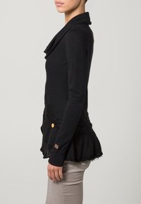 Schwarzer Pullover mit Wasserfallausschnitt, Rippdetails und Schößchen-Saum. Mit zwei Taschen und einem goldenen Knopf als Akzent auf der Vorderseite.