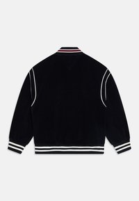 Czarna aksamitna kurtka bomberka z białymi i czerwonymi paskami na kołnierzu i mankietach, o luźnym kroju i gładkiej fakturze.