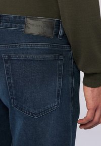 Pantalones vaqueros de mezclilla azul oscuro con un bolsillo trasero que tiene una etiqueta de cuero negro de la marca Trussardi, llevados con un suéter de manga larga verde oliva, mano visible al costado.