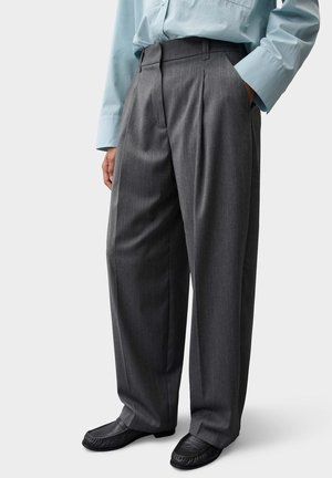 Person trägt lockere dunkelgraue plissierte Hose mit schwarzen Schuhen und hellblauem Langarmhemd, eine Hand in der Tasche, steht vor weißem Hintergrund.