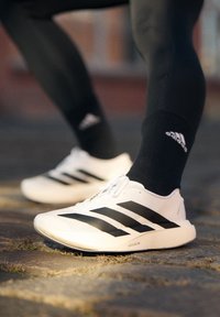 Personne portant des chaussettes Adidas noires et des chaussures de course blanches Adidas avec des rayures noires, debout sur un sol pavé.