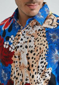 Chemise à manches courtes bleue et beige avec des motifs floraux et animaliers. Dotée de boutons et d'un col pointu, fabriquée dans un tissu lisse.